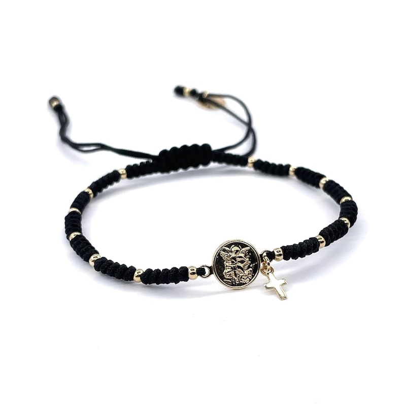 May include: A black string bracelet with gold beads and a gold charm featuring a religious image. The charm is attached to a small gold cross.