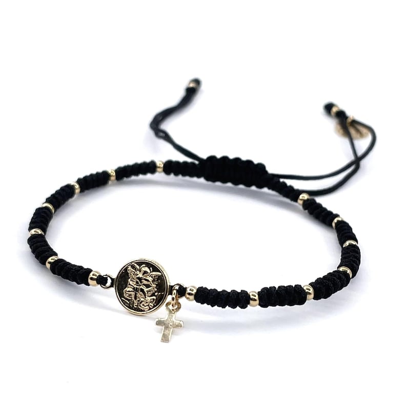 May include: Black braided bracelet with a gold charm featuring a religious icon and a small cross.