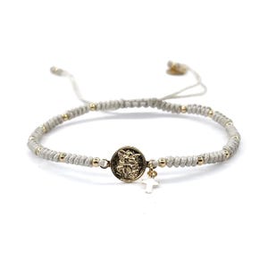 May include: A white string bracelet with gold beads and a gold charm featuring a religious image. A small silver cross charm hangs from the bottom of the gold charm.