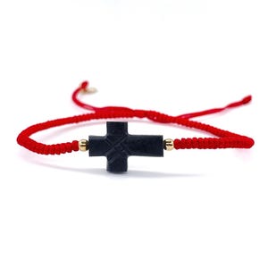 May include: A red string bracelet with a black cross charm. The cross is carved with intricate details and has a gold accent.