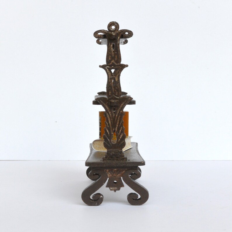 May include: A dark brown metal bookshelf with ornate scrollwork and three shelves. The bookshelf is designed to hold small books or other decorative items.