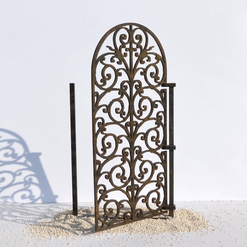 Puede incluir: Una puerta de metal decorativa con un dise&ntilde;o de desplazamiento. La puerta est&aacute; hecha de metal marr&oacute;n y tiene un aspecto r&uacute;stico. La puerta est&aacute; abierta y est&aacute; de pie sobre una superficie blanca.