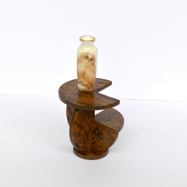 Puede incluir: Un peque&ntilde;o jarr&oacute;n de cer&aacute;mica blanca con un dise&ntilde;o de b&uacute;ho se encuentra sobre un soporte de madera marr&oacute;n de varios niveles. El soporte est&aacute; hecho de tres piezas semicirculares.