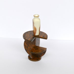 Puede incluir: Un peque&ntilde;o jarr&oacute;n de cer&aacute;mica blanco y marr&oacute;n se encuentra sobre un soporte de madera de varios niveles. El soporte est&aacute; hecho de tres piezas circulares de madera, cada una con un patr&oacute;n diferente.