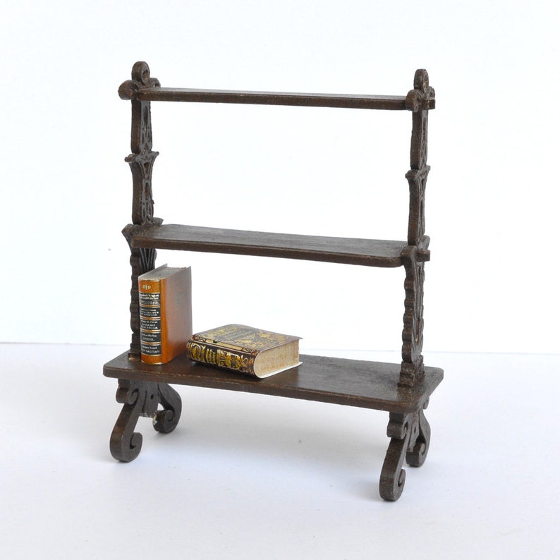 May include: A miniature wooden bookshelf with three shelves and ornate details. Two small books are displayed on the shelves.