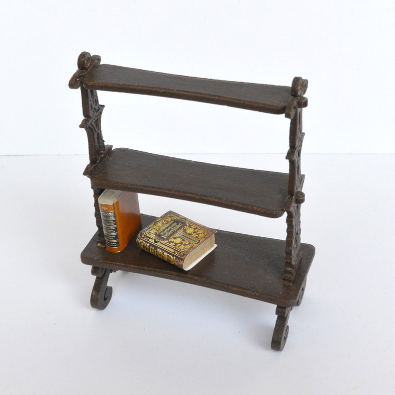 May include: A miniature wooden bookshelf with two shelves and ornate details. Two small books are displayed on the bottom shelf.