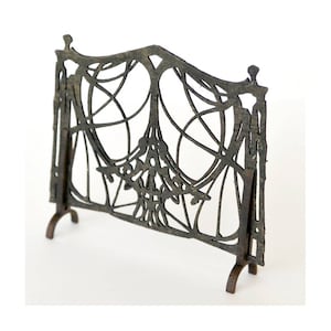 May include: A black metal fireplace screen with a decorative design of a web or lattice pattern. The screen has three legs and stands on a white surface.