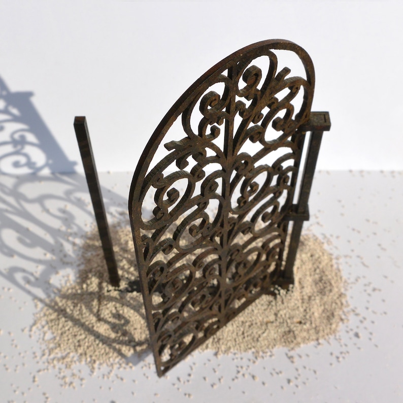 Puede incluir: Una puerta de metal decorativa con un dise&ntilde;o detallado e intrincado. La puerta est&aacute; hecha de metal marr&oacute;n y tiene un aspecto r&uacute;stico y envejecido. La puerta est&aacute; tumbada sobre una superficie blanca.