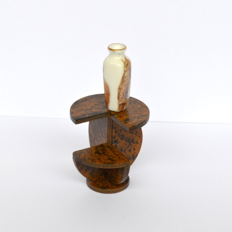 Puede incluir: Un peque&ntilde;o soporte de madera marr&oacute;n de tres niveles con un jarr&oacute;n de cer&aacute;mica blanco y marr&oacute;n en la parte superior. El soporte tiene una base circular y tres niveles.