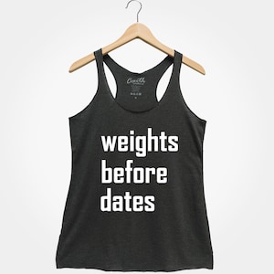 May include: Dark gray racerback tank top with the text "weights before dates" in white block letters. The tank top is hanging on a wooden hanger against a white background. The tank top is made of a soft material.