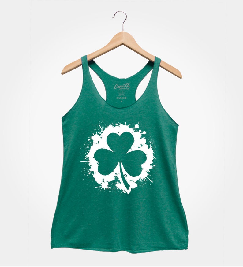 May include: A green tank top with a white shamrock graphic in a white paint splatter design. The tank top has a racerback style.