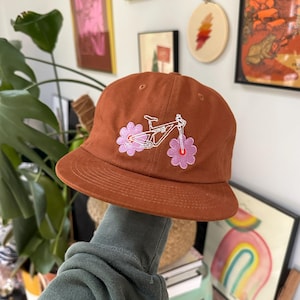 May include: A terracotta-coloured baseball cap with an embroidered bicycle design and pink flower accents. The cap has a curved peak and a classic six-panel design. This hat is a stylish accessory for outdoor activities or casual wear.