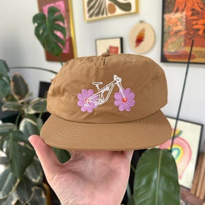 May include: A brown baseball cap featuring an embroidered bicycle design with pink flower accents. The cap has a curved brim and a rounded crown. The design is centered on the front of the cap.