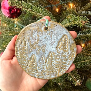 May include: A round, white and brown ceramic ornament with a design of a snowy forest scene. The ornament has a hole at the top for hanging.