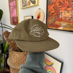 May include: A green canvas baseball cap with white embroidered text that reads "Leave it Better Than You Found It".