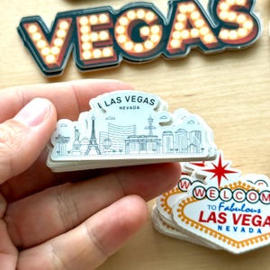 May include: A stack of white Las Vegas souvenir stickers. One sticker features a skyline with the Eiffel Tower and the text "LAS VEGAS NEVADA". Other stickers include the word "VEGAS" in marquee lights and a "Welcome to Fabulous Las Vegas Nevada" sign.