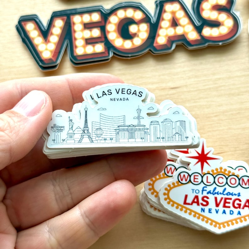 May include: A stack of white Las Vegas souvenir stickers. One sticker features a skyline with the Eiffel Tower and the text "LAS VEGAS NEVADA". Other stickers include the word "VEGAS" in marquee lights and a "Welcome to Fabulous Las Vegas Nevada" sign.