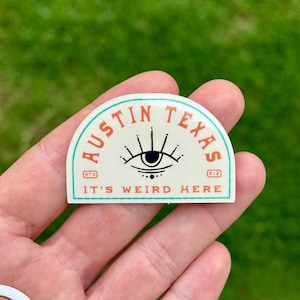 May include: A cream-colored sticker with a teal border and the words "AUSTIN TEXAS" in orange. The sticker features an eye design and the text "IT'S WEIRD HERE". The sticker also has the numbers "512" and the letters "ATX".