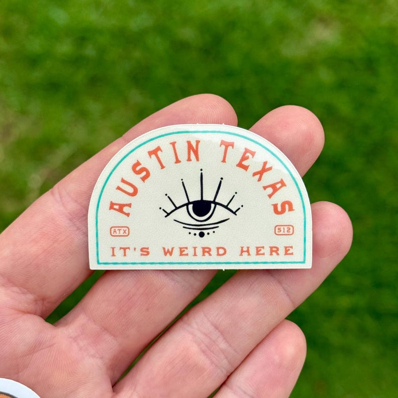 May include: A cream-colored sticker with a teal border and the words "AUSTIN TEXAS" in orange. The sticker features an eye design and the text "IT'S WEIRD HERE". The sticker also has the numbers "512" and the letters "ATX".