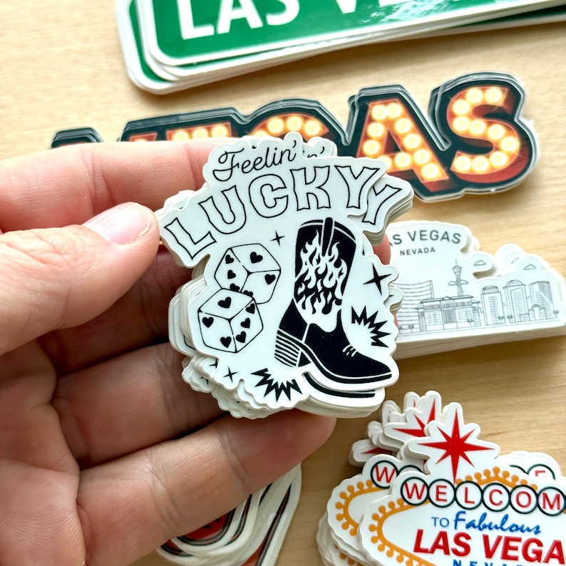 May include: A stack of white stickers with a black and white design. The main sticker says "Feelin' Lucky" with dice and a cowboy boot graphic. Other stickers include a Las Vegas sign and a "Welcome to Fabulous Las Vegas" sign.