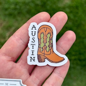 May include: A sticker featuring an orange cowboy boot with green accents. The word "AUSTIN" is printed vertically in black letters on the left side of the boot. The sticker is on a white background.