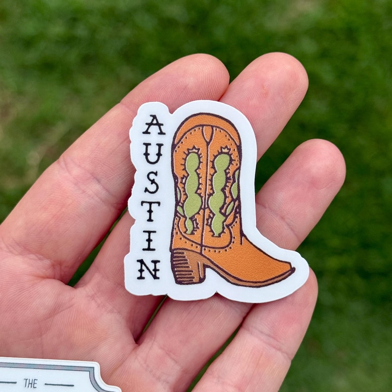 May include: A sticker featuring an orange cowboy boot with green accents. The word "AUSTIN" is printed vertically in black letters on the left side of the boot. The sticker is on a white background.