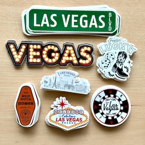 May include: A collection of Las Vegas-themed stickers. Includes a green street sign, a marquee-style "VEGAS" sign, a "Feelin' Lucky" sticker with dice and a cowboy boot, and a hotel key tag. Other stickers feature the Las Vegas skyline and a casino chip.