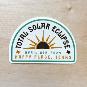 May include: A white sticker with a green border in the shape of a half circle. The sticker has a black and gold sun graphic with the text "TOTAL SOLAR ECLIPSE" in black lettering. The text "APRIL 8TH. 2024" and "HAPPY PLACE, TEXAS" are also on the sticker.