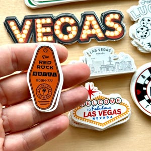 May include: A collection of Las Vegas-themed stickers. One sticker is an orange hotel key tag that reads "The Red Rock Hotel, Room-777". Other stickers include the word "VEGAS" in marquee lights and a "Welcome to Fabulous Las Vegas" sign.