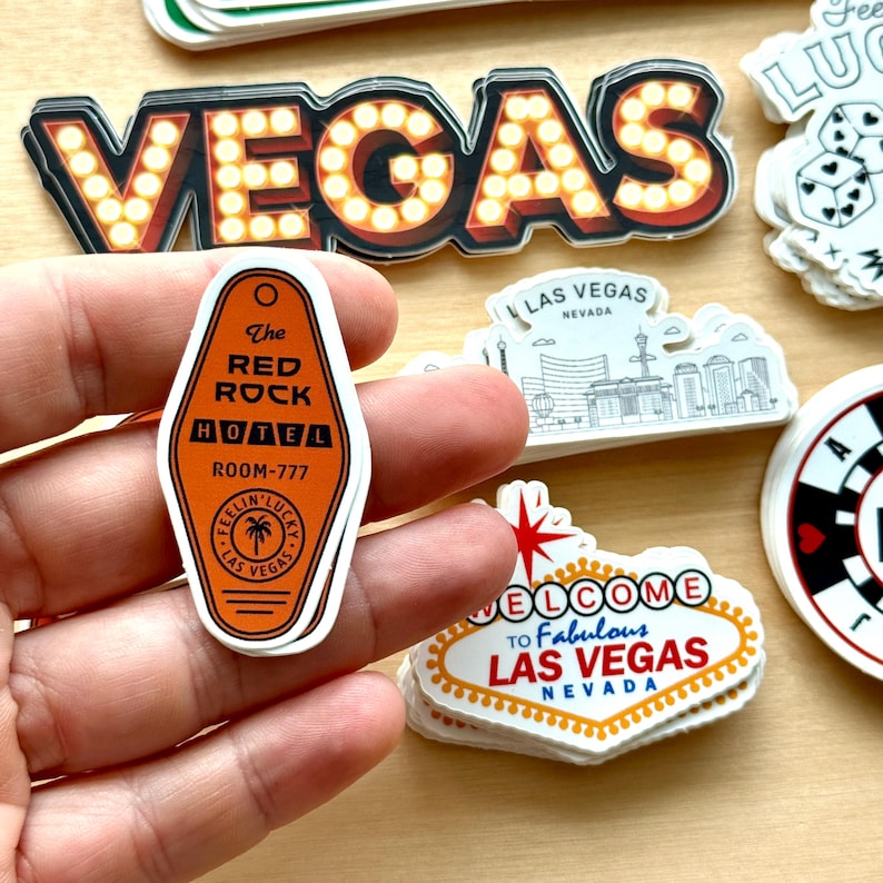 May include: A collection of Las Vegas-themed stickers. One sticker is an orange hotel key tag that reads "The Red Rock Hotel, Room-777". Other stickers include the word "VEGAS" in marquee lights and a "Welcome to Fabulous Las Vegas" sign.