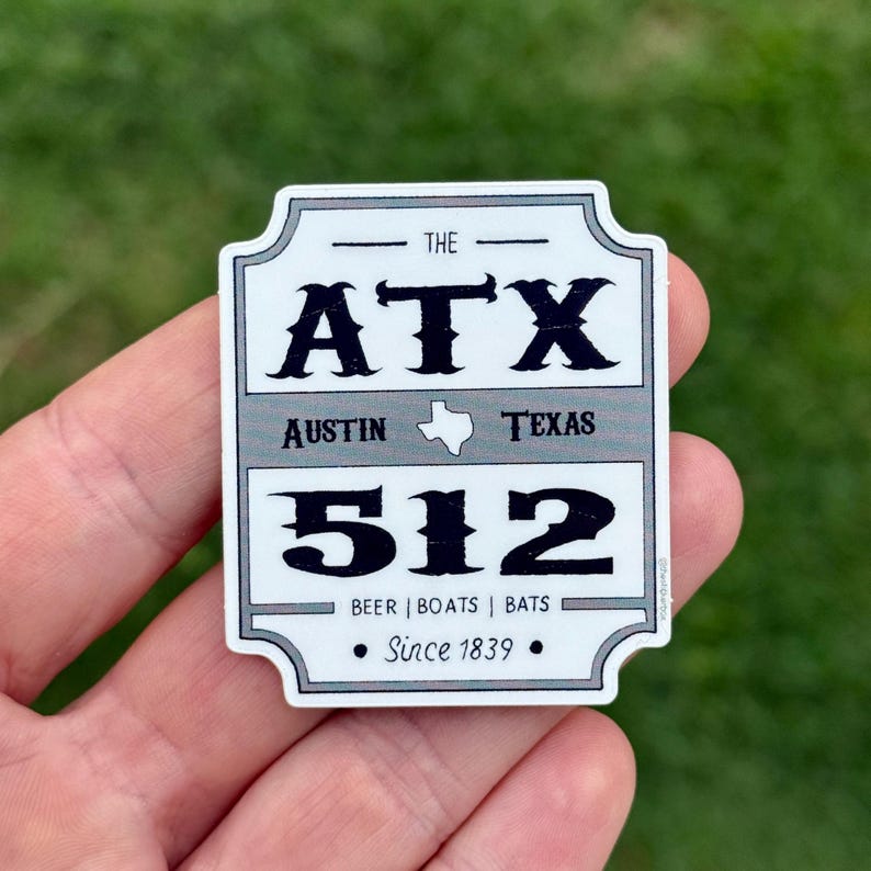May include: A rectangular sticker with a gray and white design. The sticker reads "THE ATX AUSTIN TEXAS 512" with the words "BEER | BOATS | BATS" and "Since 1839" at the bottom. The sticker is held in a person's hand.