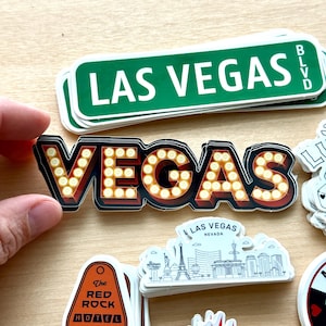 May include: A collection of Las Vegas-themed stickers. One sticker features the word "VEGAS" in a marquee style with illuminated bulbs. Other stickers include a green street sign that reads "LAS VEGAS BLVD" and a Red Rock Hotel sticker.