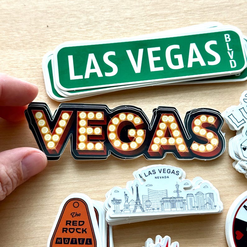 May include: A collection of Las Vegas-themed stickers. One sticker features the word "VEGAS" in a marquee style with illuminated bulbs. Other stickers include a green street sign that reads "LAS VEGAS BLVD" and a Red Rock Hotel sticker.