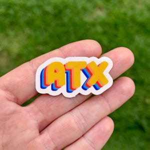 May include: A colorful sticker with the letters "ATX" in a 3D effect. The letters are yellow with red and blue outlines, set against a white background. The sticker is held in a hand against a green background.
