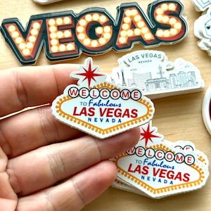 May include: A collection of Las Vegas-themed stickers. One sticker features the iconic "Welcome to Fabulous Las Vegas" sign with a red star and the text "Welcome to Fabulous Las Vegas Nevada". Other stickers include the word "VEGAS" with illuminated letters and a Las Vegas skyline.