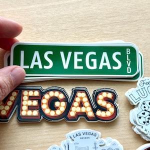 May include: A collection of Las Vegas-themed stickers. One sticker is a green street sign that reads "LAS VEGAS BLVD" in white. Another sticker spells out "VEGAS" in illuminated marquee-style letters. Other stickers are also visible.
