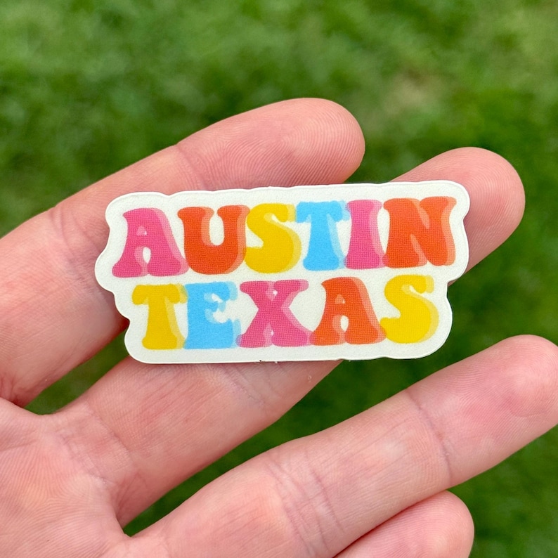 May include: A colorful sticker with the words "AUSTIN TEXAS" in a retro font. The text is in shades of pink, orange, yellow, and blue, with a white border. The sticker is held in a hand against a blurred green background.