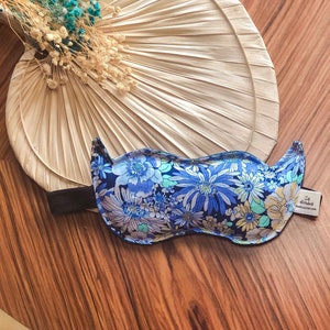 May include: A blue and white floral eye mask with a black elastic strap. The eye mask is on a wooden table with a fan-shaped object in the background.