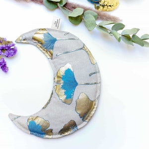 May include: A crescent moon-shaped heating pad with a light gray fabric and a blue and gold ginkgo leaf pattern. The pad is on a white background.