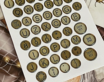 May include: A set of gold-coloured typewriter keys with black lettering. The keys are arranged in a keyboard layout and include letters, numbers, symbols, and punctuation marks. The keys are on a white background.