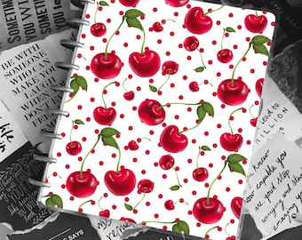 May include: A white planner with a red cherry and white polka dot pattern. The planner has a clear plastic cover and a metal ring binding. The planner is surrounded by black and white paper scraps with inspirational quotes.