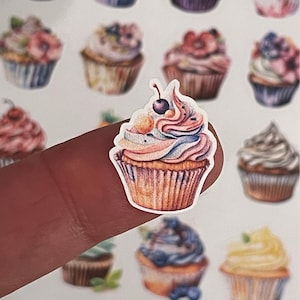 以下が含まれることがあります： ピンク、ブルー、イエローのフロスティングと、上に赤いチェリーが1つ乗ったカラフルなカップケーキのステッカー。ステッカーは指の上にあり、背景にはたくさんのカップケーキのステッカーがあります。