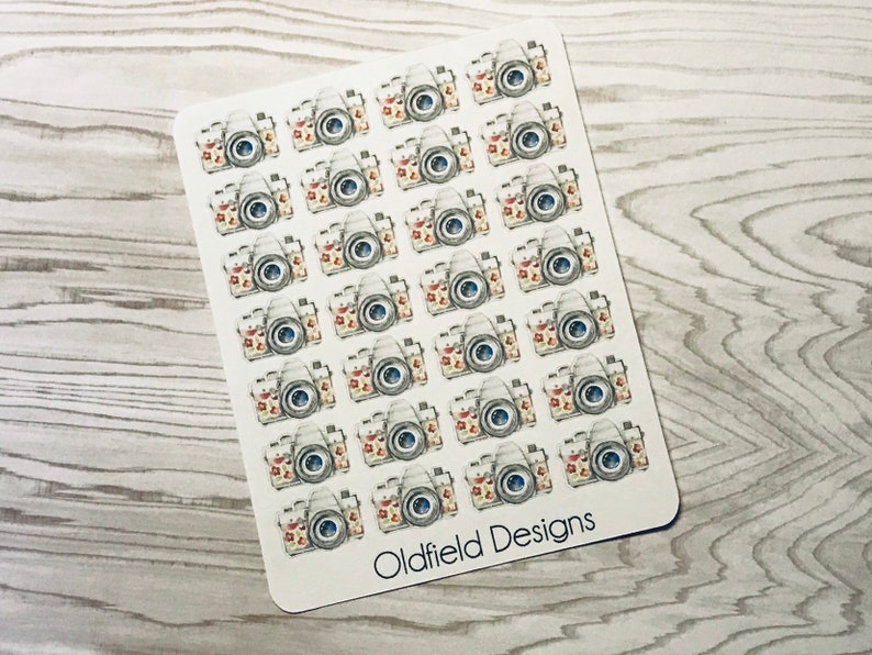 Pu&ograve; includere: Un foglio di adesivi con fotocamere in stile acquerello con accenti floreali. Le fotocamere sono principalmente grigie con dettagli rossi, gialli e blu. Il testo "Oldfield Designs" &egrave; stampato in basso.