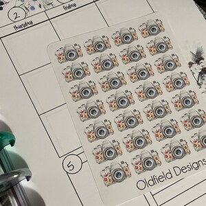 Pu&ograve; includere: Foglio di adesivi con fotocamere ad acquerello con accenti floreali. Le fotocamere hanno corpi grigi, lenti blu e motivi floreali colorati. Gli adesivi sono su sfondo bianco con una pagina di pianificatore visibile. Il testo "Oldfield Designs" &egrave; visibile.