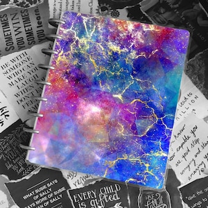 Op de afbeelding: Een kleurrijk notitieboek met een galaxy-achtig ontwerp en gouden accenten. Het notitieboek heeft een spiraalbinding en is omringd door inspirerende citaten.