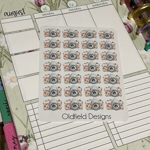 Pu&ograve; includere: Un foglio di adesivi con fotocamere ad acquerello con accenti floreali. Gli adesivi sono disposti su uno sfondo bianco, con il testo "Oldfield Designs" in basso. Gli adesivi sono su un'agenda con il mese di agosto visibile.