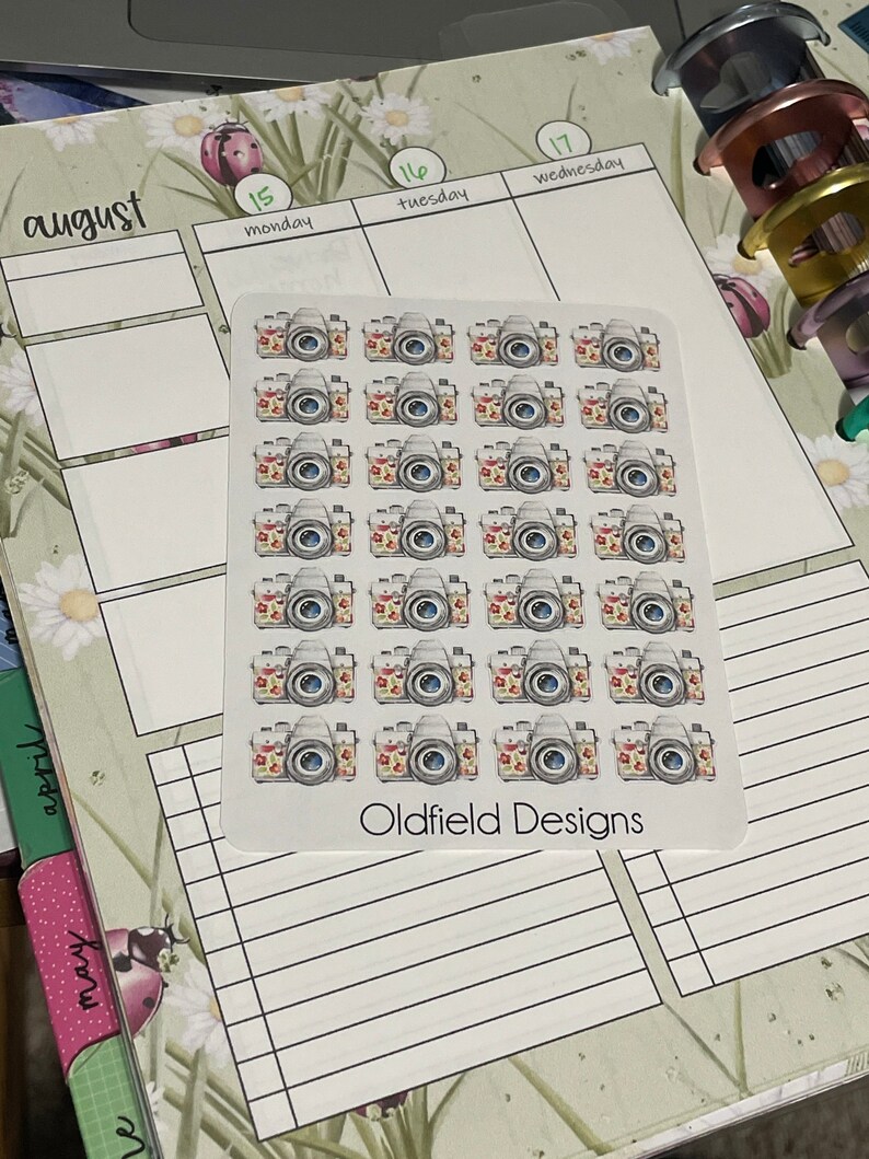 Pu&ograve; includere: Un foglio di adesivi con fotocamere ad acquerello con accenti floreali. Gli adesivi sono disposti su uno sfondo bianco, con il testo "Oldfield Designs" in basso. Gli adesivi sono su un'agenda con il mese di agosto visibile.