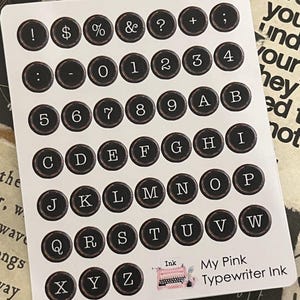 May include: A sheet of typewriter key stickers with black keys and white lettering. The keys include numbers, letters, and symbols. The text "My Pink Typewriter Ink" and a pink typewriter graphic are in the bottom right corner.