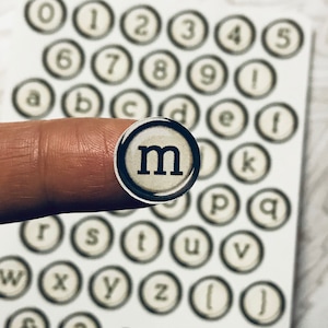 May include: A set of typewriter key stickers with letters, numbers, and symbols. The stickers are white with black lettering and are arranged in rows on a white background. The letter "m" is highlighted in the image.