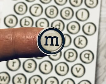 Può includere: Un set di adesivi per tasti di macchina da scrivere con lettere, numeri e simboli. Gli adesivi sono bianchi con lettere nere e sono disposti in file su uno sfondo bianco. La lettera "m" è evidenziata nell'immagine.